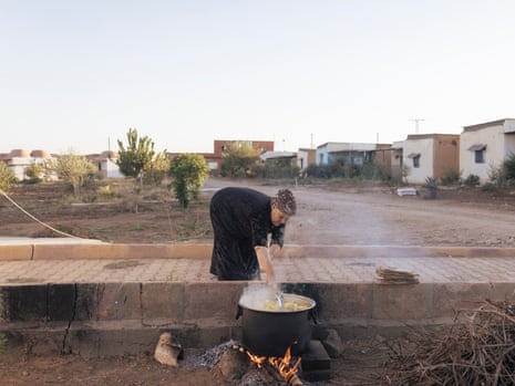 A woman stands on a road bending over a large cooking pot on an open fire