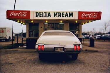 A white car heading into a drive-in restaurant with the words ‘Delta Kream’ and ‘Coca-Cola’ above the ordering window