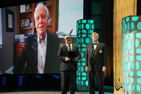 A an older man's image is projected behind two other men in formalwear on stage.
