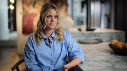 she is in her early 30s and has long blond, wavy hair; she wears a pale blue cotton shirt and is sitting in a chair in a luxurious-looking room with a painting behind her.