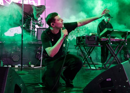 A man kneels down and reaches out one arm and holds a mic with the other while performing on a stage filled with green smoke.