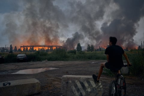 A teenager sits on a bicycle watching flames rise from a burning logistics warehouse.