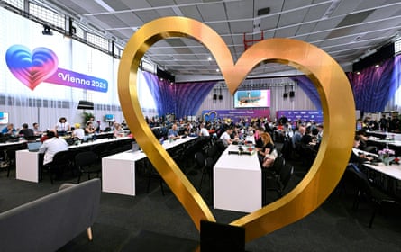 Large gold heart logo in foreground with Vienna 2026 sign to left with journalists worling behind