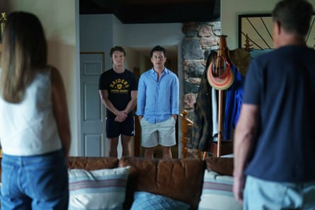 Two men stand in a cottage doorway looking ahead, past sofa cushions and other people