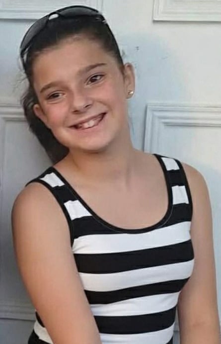 she has light brown hair and wears a black and white striped vest top. She has long light brown hair which is tied back, and wears a pair of sunglasses pushed back on the top of her head. She is smiling in this photo.