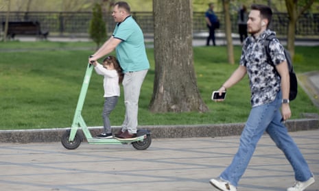 A young girl shares a green scooter with a man, and together they ride it on the pavement next to the green lawn of Volodymyrska Hill Park in Kyiv. A man walks near the pair on the same pavement. 