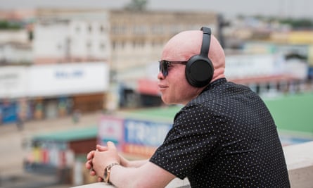 A man wearing a headphones and sunglasses leans on a wall and looks out over a city