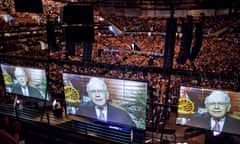 A covered stadium full of people, with multiple, giant side-by-side screens of an older white man in a suit speaking from what appears to be an office.