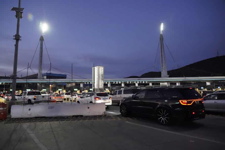 Cars approach a barrier between the US and Mexico at dusk.