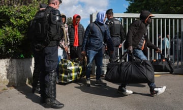 Gendarmes oversee migrants being evacuated from a squat in a disused factory: young men, mostly black and wearing anoraks and hooded tops, walk out of a gate carrying large bags as police look on.