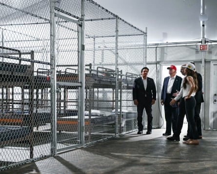 Donald Trump, Ron DeSantis, Kristi Noem and a guide stand in a room looking at a cage filled with bunch beds.
