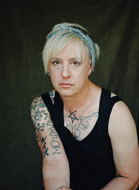 A portrait of a woman with blond hair and tattoos on her right arm looking at camera