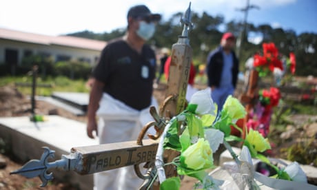 Flowers and a cross on a grave with mourners seen in the background