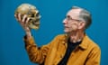Svante Pääbo, with metal-framed glasses and wearing a button-down shirt over a T-shirt, looks at the model of a Neanderthal skull he is holding up