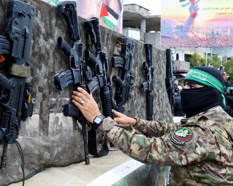 A Hamas militant in camouflage and a green headband reaches toward assault rifles mounted on a mesh display