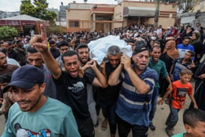 Covered in a white sheet, a body is carried on the shoulders of mourners through a crowded street