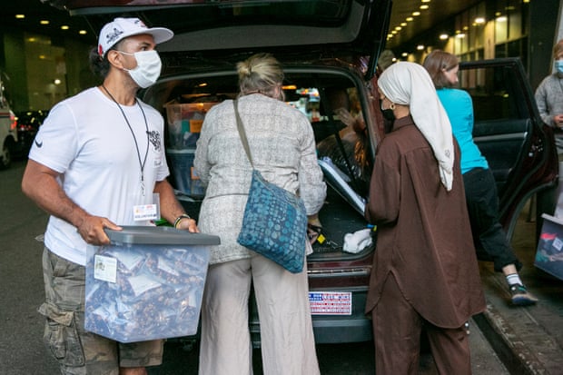 A man wearing a face mask unloads a clear plastic tub from the back of a car as two women peer into the trunk space, looking at other supplies.