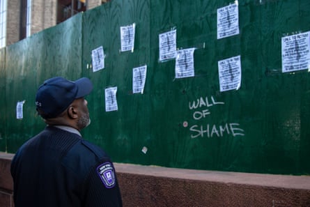 campus security officer in blue baseball cap and jacket looks at wall made of wooden board, featuring small posters saying ‘call to action’, with the words ‘wall of shame’ written under the posters