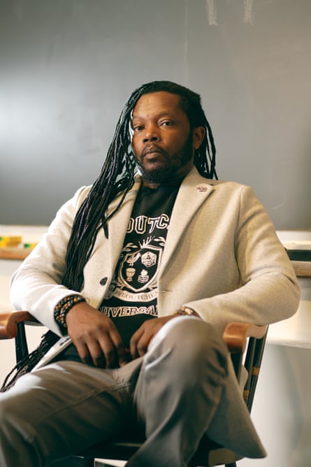 Black man with very long dreads, wearing black T-shirt, cream blazer, and gray khakis leans back in chair in classroom, looking seriously at camera.