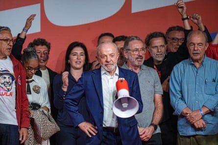 Lula stands on a stage holding a red and white megaphone, looking sombre. People behind him hold their hands and also look subdued, with only a few raising their arms.
