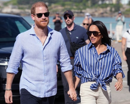 Prince Harry and Meghan Markle walk hand in hand, both wearing blue linen shirts, on a visit to Bondi Beach in Australia in April
