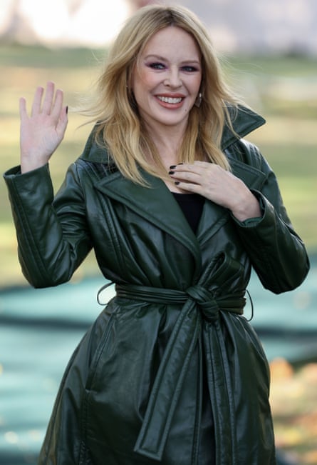 Kylie Minogue, with long hair, in a green trench coat waves with her right hand and touches her collarbone with her left hand