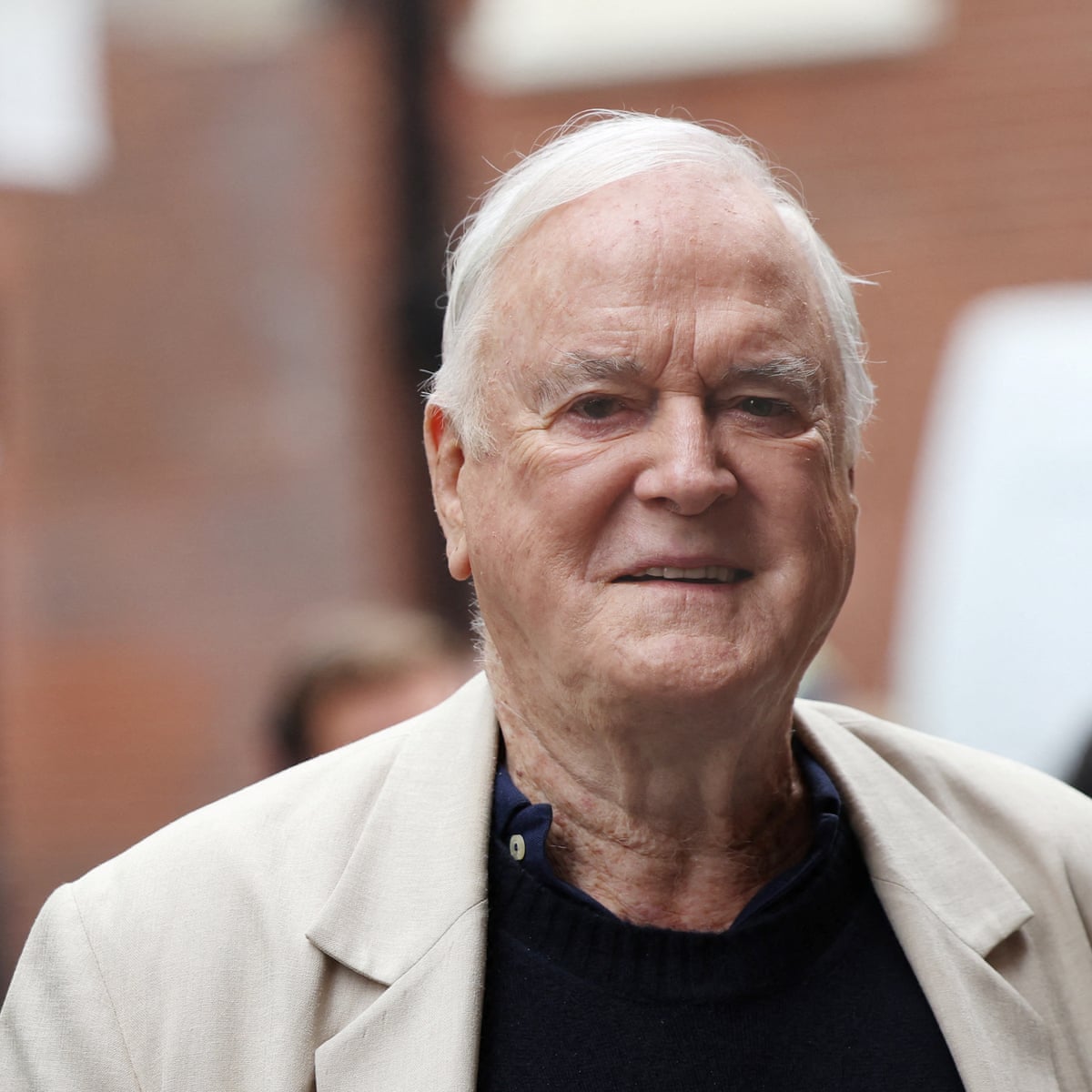 John Cleese, Monty Python comedian and actor, wearing a light jacket and dark shirt while smiling outdoors.