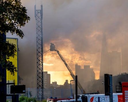 A firefighter on a crane directs a fire hose seen against smoke filling the London skyline backlit by sunset