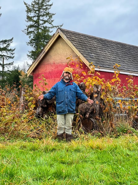 Zach Galifianakis wearing a blue jacket, grey trousers and a tan trappers hat stands with his arms outstretched in a field by a red painted barn