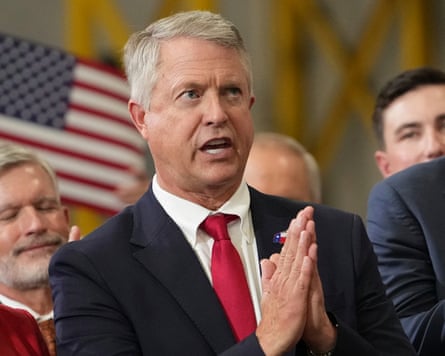 An older man in a suit in front of a US flag.