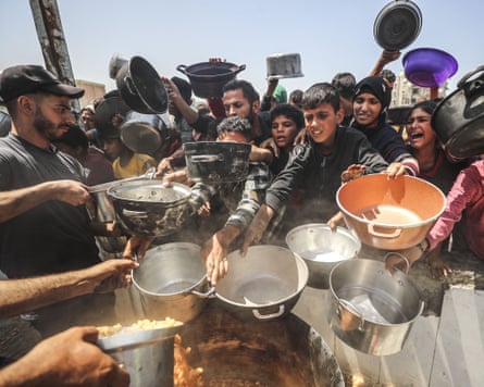 A group of Palestinians crowd around a large pot with bowls, appealing for food.