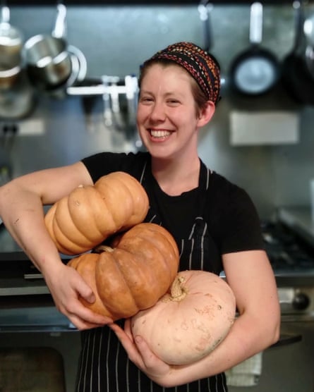 A chef, Lucy Ridge, in a black top and apron, smiling in a commercial kitchen and holding three large pumpkins.
