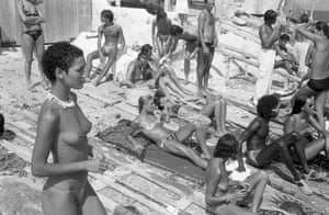 Black-and-white photo of men and women standing and lying on a beach. Most of the women are topless