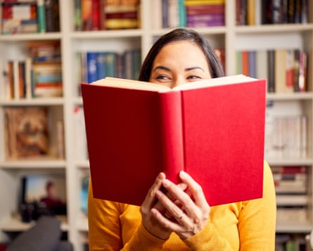 Smiling beautiful young female reading book standing against bookshelf at home