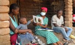 An African woman in a facemask sits with a young couple and their baby on the steps outside a house