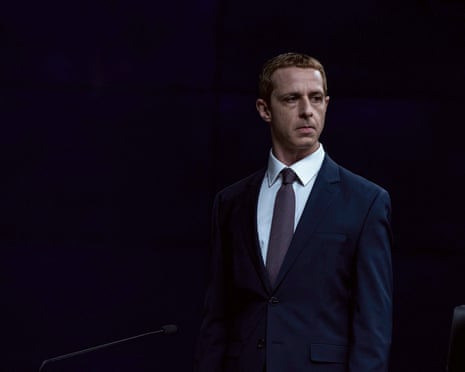 A serious looking man wears a dark blue suit and plain dark tie, standing against a black background