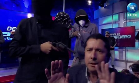 A man in a TV studio holds his hands in the air as two gunmen wearing masks and balaclavas point guns at his head.