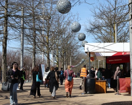 People walk through a paved park with trees and stalls