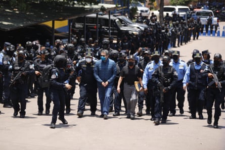 A handcuffed man with a face mask is surrounded by armed police, also in face masks