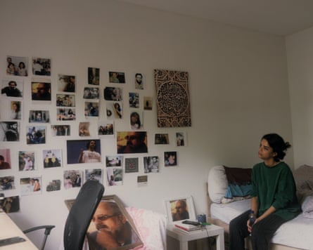 A woman sites on her bed looking longingly at photos on the wall.