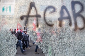 Protestors pass a smashed bus stop with graffiti reading ACAB ( All coppers are bastards) 