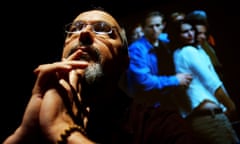 Bill Viola, with his hands on his chin, seen from below in a dark room, with a video showing a crowd behind him
