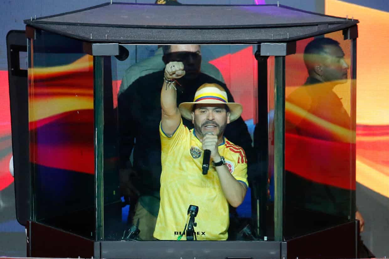 Abelardo de la Espriella raises a fist as he speaks into a microphone while standing in a protective glass enclosure