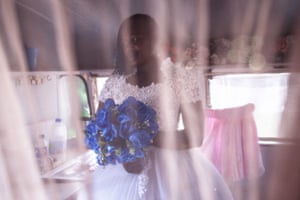 Memory poses in her dress with blue bouquet, shot through a net curtain inside the caravan