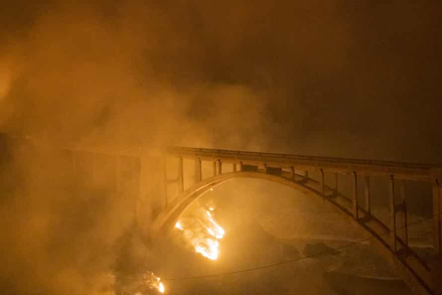 A reinforced concrete arch bridge is nearly obscured by smoke as a fire burns below.