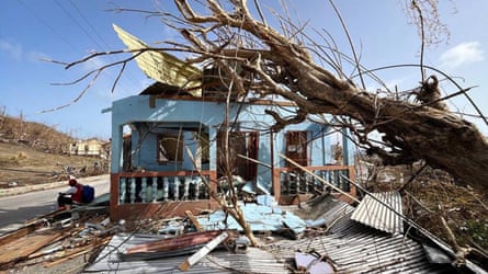 A man sits by the side of the road next to a damaged house and tree that has fallen on top of it