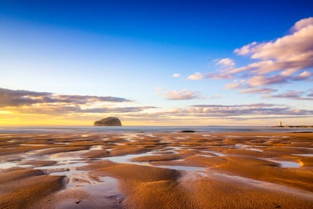 A large rock island, seen from a beach with channels of water through the sand