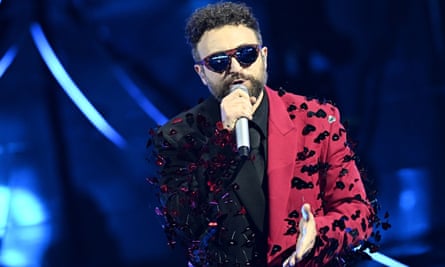  he has a beard and is wearing big sunglasses and a black shirt and tie with a half black, half crimson jacket which is covered in large sequined hearts