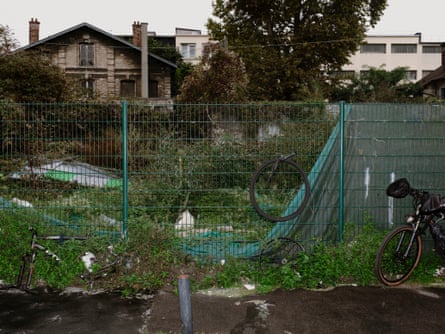 A patch of overgrown wasteland, fenced off and strewn with rubbish, behind a house and flats