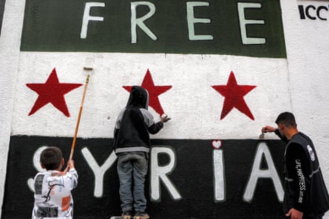 A man and two children painting a large mural that bears the words: ‘Free Syria’.